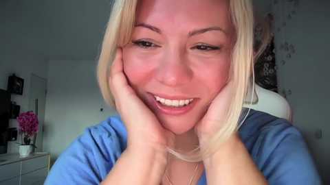 Media: A smiling, light-skinned blonde woman with shoulder-length hair, wearing a blue shirt, sits with hands supporting her face, in a softly lit room with floral decor.