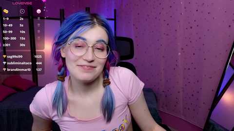 Media: Video of a fair-skinned woman with blue hair in twin braids, wearing glasses, pink t-shirt, and a playful smile, in a pink bedroom with purple lighting.