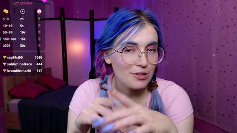 Media: Video of a young woman with vibrant blue hair, wearing large round glasses, and light pink top, sitting in a room with a bed, purple walls, and social media app interface in the corner.