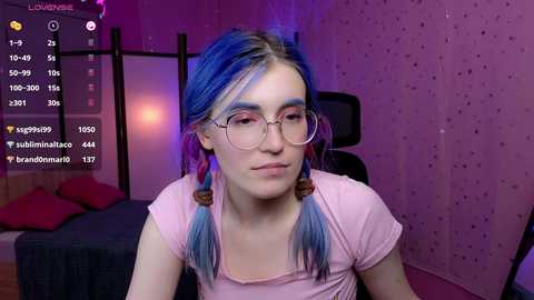 Media: Video of a young woman with blue hair in twin braids, wearing glasses and a pink t-shirt, sitting in a dimly lit room with pink walls and a bed in the background.