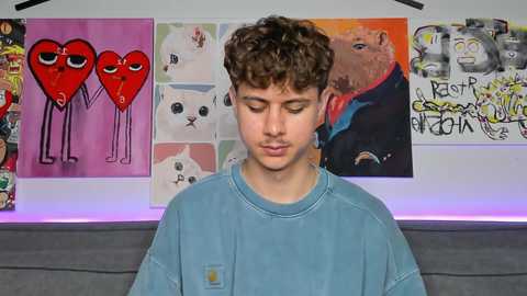 Media: Video of a young man with curly brown hair, wearing a blue shirt, standing in front of a wall adorned with colorful, abstract, graffiti-style art.