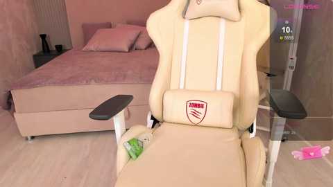 Media: Video of a beige gaming chair with a small red and black logo, in a pink and beige bedroom with a neatly made bed, a medical device, and a pink toy on the floor.