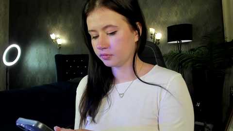 Media: Video of a young woman with fair skin and straight dark hair, wearing a white long-sleeve shirt, sitting in a dimly lit room with a black chair, a lamp, and a ring light.