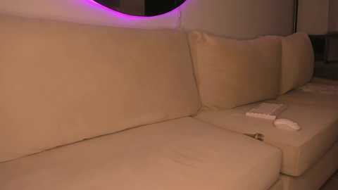 Media: Video of a beige sectional sofa in a dimly lit room, with a purple LED strip lighting the top edge. Two white remote controls are placed on the right side of the sofa.