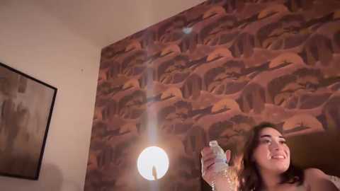 Media: A video of a young woman with dark hair, smiling while drinking from a water bottle, set against a wall with a patterned, earth-toned wallpaper depicting elephants.