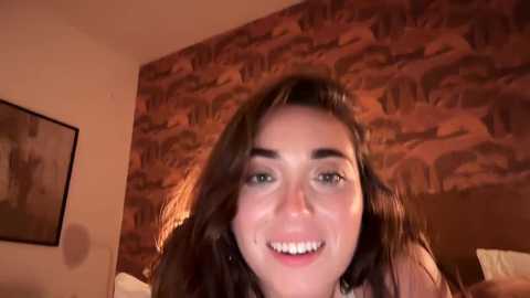 Media: Video of a smiling woman with long brown hair, fair skin, and green eyes, sitting on a bed with a brown and beige patterned wall behind her.