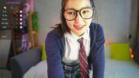 Media: Video of a smiling Asian woman with glasses, wearing a school uniform, leaning forward in a cozy bedroom with colorful decor.