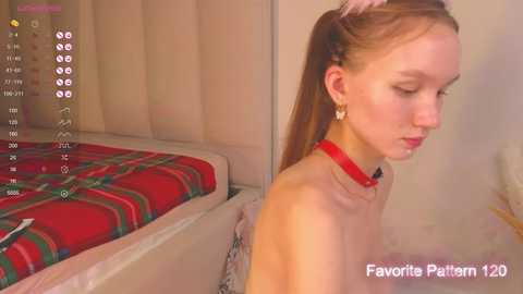 Media: Video of a young, fair-skinned woman with long brown hair in a ponytail, wearing a red choker and a white top, sitting on a bed with a red plaid blanket.