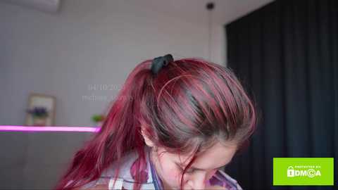 Media: Video of a young woman with long, vibrant red hair in a high ponytail, wearing a colorful, patterned shirt, in a modern, minimalist room with a pink LED strip and dark curtains.