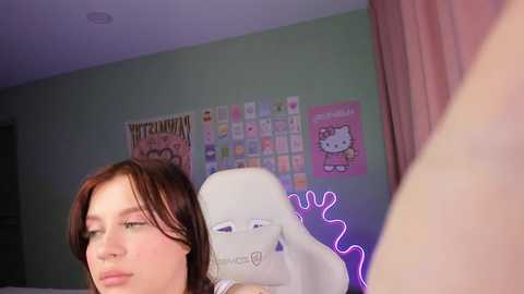 Media: Video of a young woman with long, wavy brown hair and pale skin, sitting on a white chair in a brightly lit room. Behind her, a pink Hello Kitty poster and a wall of colorful stickers adorn the wall.