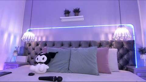 Media: Video of a modern bedroom with a plush, tufted gray headboard, two potted plants on shelves, two crystal chandeliers, and a plush white toy with black glasses on a bed.