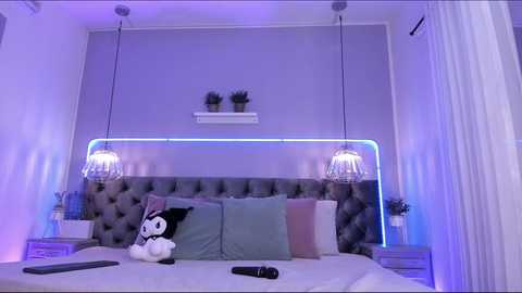 Media: Video of a modern, stylish bedroom with a plush gray tufted headboard, two potted plants, and two hanging pendant lights. The room has a purple and blue color scheme.