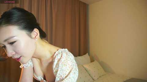 Media: A video of an East Asian woman with fair skin, black hair in a bun, wearing a floral dress, smiling, in a beige room with brown curtains and a bed with white pillows.