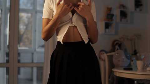 Media: Video of a light-skinned woman with medium-length hair, wearing a white crop top and black pleated skirt, standing indoors with a mirror, shelves, and a window in the background.