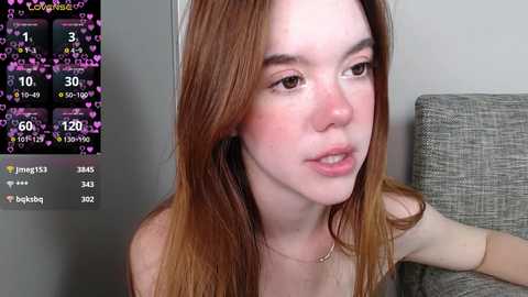 Media: Video of a young woman with fair skin and long, straight, auburn hair, wearing a light-colored top. She sits on a grey couch, with a digital screen displaying chat hearts and numbers in the background.