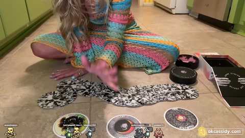 Media: Video of a woman in a tie-dye dress, sitting on a tiled floor, playing a board game. The background features a kitchen with green cabinets.