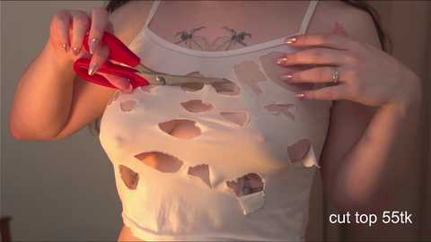 Media: A video of a woman with fair skin, wearing a white, cut-out top, using a red glue gun to apply adhesive to the garment. Her nails are manicured, and she has a tattoo on her chest.