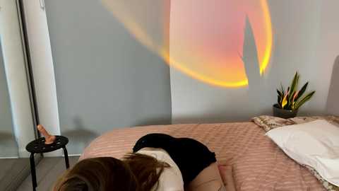 Media: Video of a woman in a white shirt and black shorts lying on a pink quilted bed, with a colorful light projection on the wall behind her.