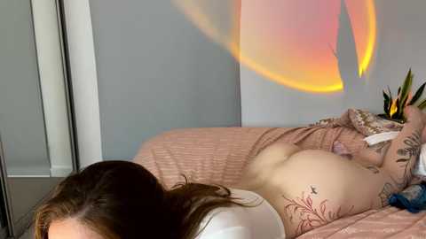 Media: Video of a nude woman with fair skin and brown hair, lying on a pink bedspread, showcasing her tattooed back and buttocks. A colorful, abstract wall mural with a sun motif adds a vibrant backdrop.