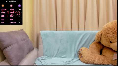 Media: A video of a cozy room featuring a beige teddy bear on a light blue blanket, with a lavender cushion on a white couch. The background has beige curtains. A digital display on the left shows a score and heart rate.