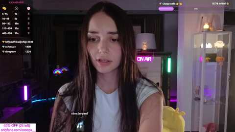 Media: Video of a young woman with long dark hair, wearing a white t-shirt and plaid vest, in a dimly-lit room with neon lights and a glass cabinet.