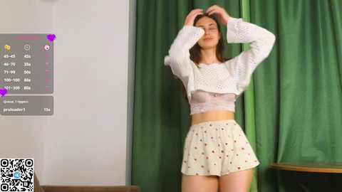 Media: Video of a young woman with light skin and long brown hair, wearing a sheer white crop top and floral skirt, standing in front of green curtains.