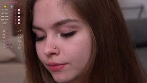 Media: Video of a young woman with fair skin, long brown hair, and a small nose piercing. She has closed eyes and a slight smile, looking down. Background is blurred.