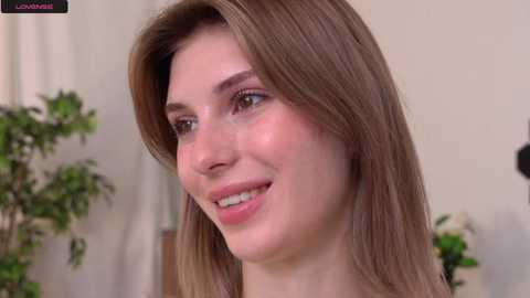 Media: Video of a smiling young woman with straight, light brown hair, fair skin, and pink lips, set against a blurred indoor background featuring green plants and white curtains.