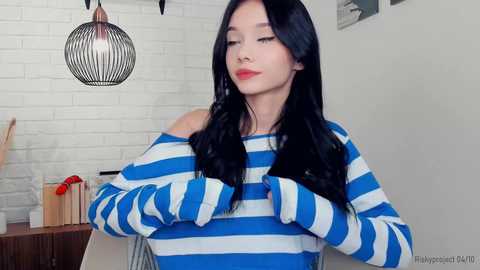 Media: Video of an East Asian woman with long black hair, wearing a blue and white striped off-shoulder top, posing indoors with a white brick wall background and modern pendant light.