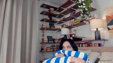 Media: Video of a woman with black hair, wearing a blue and white striped blanket, peeking out from behind a bookshelf in a cozy, cluttered living room with wooden shelves, books, and a lamp.