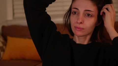 Media: Video of a young woman with fair skin and shoulder-length dark hair, wearing a black sweater, adjusting her hair, indoors with a brown couch and orange pillow in the background.