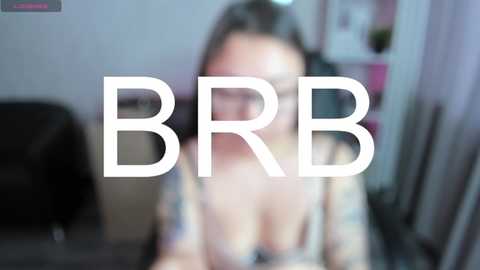 Media: Video of a blurred woman wearing glasses and a floral blouse, with text \"BBR\" overlaid. The background shows a dimly lit room with furniture and a wall.