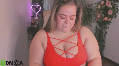 Media: Video of a plus-size, light-skinned woman with shoulder-length brown hair wearing a red crop top with crisscross straps. Background features a heart-shaped neon light and floral decorations.