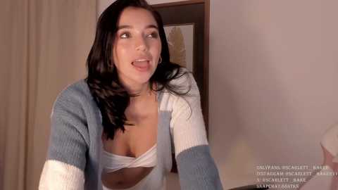 Media: Video of a young woman with long black hair, wearing a blue and white striped cardigan over a white bra, looking playful and smiling.