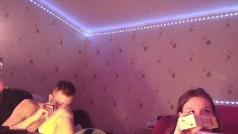 Media: Video of a dimly lit room with pink and blue LED strip lights. Two young boys, one shirtless, and a girl with dark hair sit on a bed, partially hidden by a blanket, laughing and holding their noses.