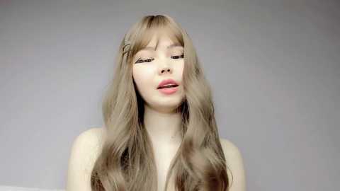 Media: Video of a young woman with long, wavy, light brown hair and fair skin. She has a neutral expression, slightly parted lips, and is topless, set against a plain gray background.