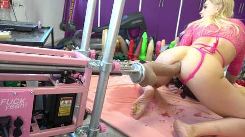 Media: Video of a blonde woman in pink lingerie kneeling on a pink mat, engaging in a sex act with a large, erect penis attached to a machine in a gym setting.