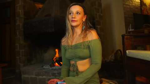 Media: Video of a blonde woman in a green off-shoulder top and skirt, sitting near a fireplace in a cozy room with brick walls and a wooden table.