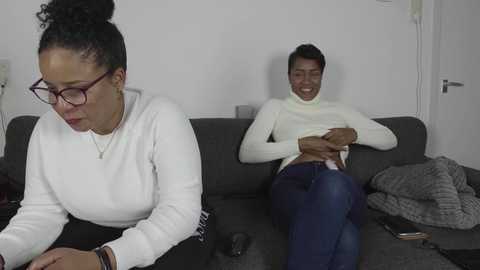 Media: Video of a dark-skinned woman with curly hair in glasses, wearing a white sweater, sitting on a gray couch, frowning, next to a dark-skinned man in a white turtleneck and blue jeans, laughing, in a minimalist living room.