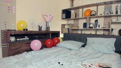 Media: Video of a cozy bedroom with a gray upholstered headboard, light blue striped bedding, a wooden dresser holding yellow and pink balloons, and a shelving unit with books, a clock, and a vase.