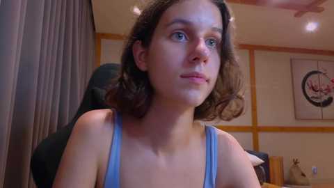 Media: Video of a young woman with wavy brown hair, light skin, and blue eyes, wearing a light blue tank top, seated indoors with beige walls, a framed circular art piece, and a wooden chair in the background.