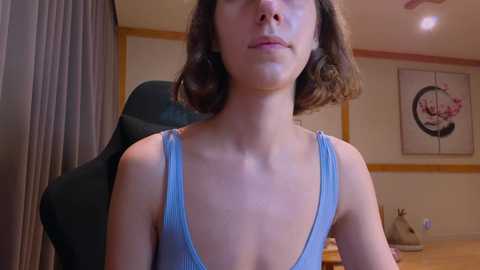 Media: Video of a young Caucasian woman with short brown hair, wearing a light blue tank top, sitting in a room with beige walls, a black chair, and a decorative painting.