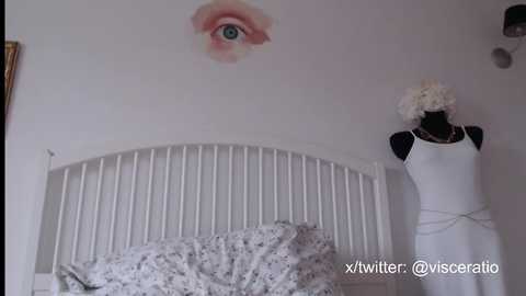 Media: Video of a white bed with a white headboard featuring a floral decal of an eye, a white dress mannequin, and a white bedspread with a subtle pattern.