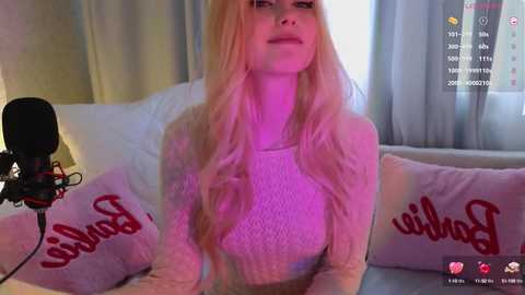 Media: A video of a fair-skinned, long-haired, blonde woman wearing a knit sweater, sitting on a bed with red pillows, under purple lighting, with a camera setup visible in the background.