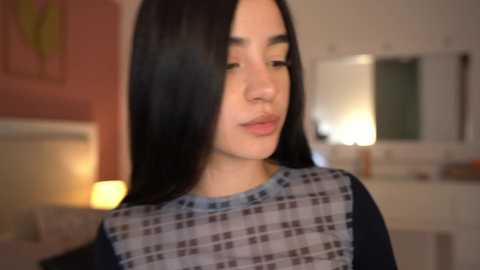 Media: A video of a young woman with long, straight black hair, fair skin, and closed eyes, wearing a blue and gray checkered apron, standing in a softly lit, cozy kitchen with warm tones and modern decor.