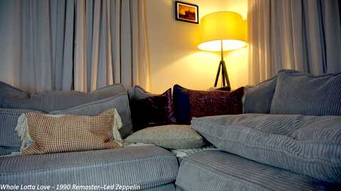 Media: Video of a cozy living room with a large, plush grey sectional sofa adorned with striped and checkered cushions. A warm yellow lamp with a pleated shade casts a soft glow against grey curtains.