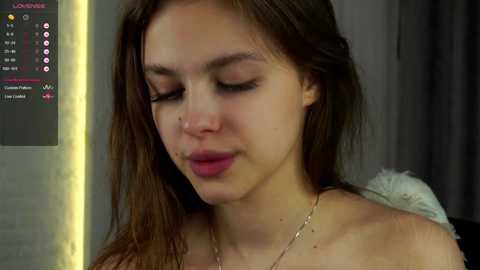 Media: Video of a young woman with fair skin, brown hair, and a slight nose piercing, wearing a delicate necklace. She appears contemplative with a neutral expression, indoors against a gray curtain backdrop.