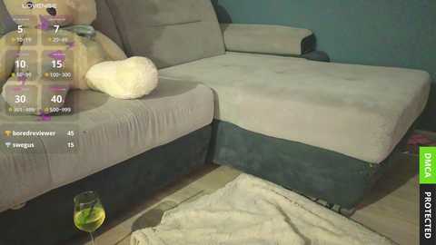 Media: Video of a beige sectional sofa with a blue base, featuring a plush white pillow and a glass of yellow liquid on the floor.