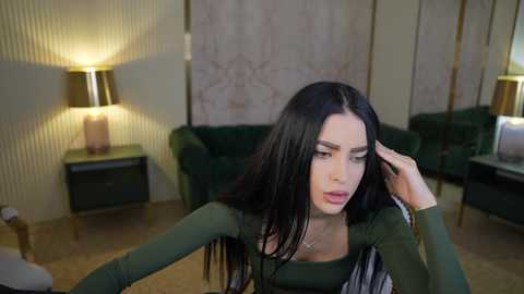 Media: Video of a woman with long black hair, wearing a green top, adjusting her hair in a modern, dimly-lit living room with green couches, beige walls, and a gold lamp.