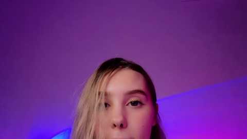 Media: Video of a young Caucasian woman with long blonde hair, wearing golden eyeshadow, captured against a purple and blue gradient background. Her expression is contemplative, with a slight tilt of her head.